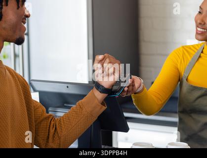 Female barista and African American male customer tapping smartwatch on reader at coffee counter Stock Photo