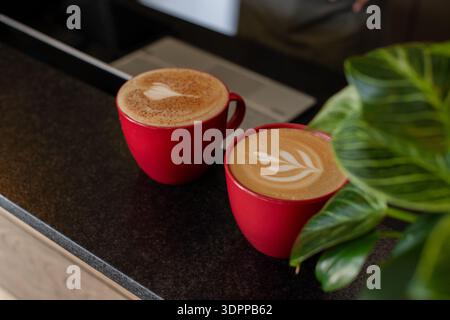 Two red mugs displaying latte art resting on stone countertop near laptop, green plant in cafe Stock Photo