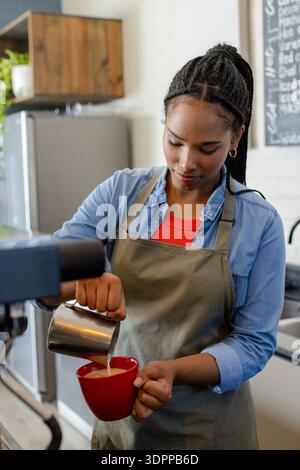 African American woman barista pouring steamed milk from steel pitcher into red mug at cafe Stock Photo