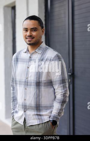 Man standing by white wall and dark grey louvered door on paved walkway while checking smartwatch Stock Photo