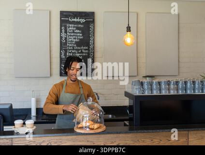 African American male barista lifting glass dome over croissants behind counter by takeaway cups Stock Photo