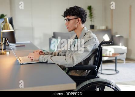Asian man using wheelchair typing on laptop at desk featuring gold lamp and monitor in office Stock Photo