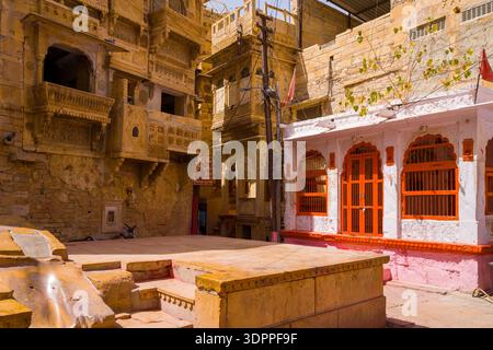 Jaisalmer, India - March 28, 2022: The image shows a sandstone courtyard surrounded by yellow sandstone buildings with ornate carved facades and balconies, and a white building with red-painted arches and metal bars in Jaisalmer. Stock Photo