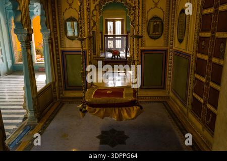 Udaipur, India - March 24, 2022: An ornately decorated room inside City Palace features a traditional royal swing suspended from the ceiling, with intricate wall designs, mirrors, and a view into adjacent chambers with striped flooring and colorful walls. Stock Photo