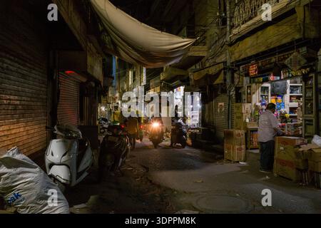 Jodhpur, India - March 25, 2022: The image shows a narrow street at night with an open shop displaying snacks and goods, a man handling boxes, parked scooters, an auto rickshaw, and people walking and riding motorcycles under overhead wires and fabric canopy. Stock Photo