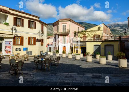 Maratea, Italy - October 10, 2020: The image shows an empty outdoor cafe area with tables and chairs arranged on a stone-paved square, surrounded by pastel-colored historic buildings and mountain scenery in the background. Stock Photo