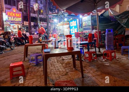 Ho Chi Minh City, Vietnam - January 13, 2020: Sidewalk food stall at night with plastic tables and stools, illuminated signage, drink refrigerator, and people on motorbikes visible in the background. Stock Photo