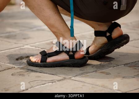 Male tourist in black strap sandals crouching on stone ground, leg skin, high angle view Stock Photo