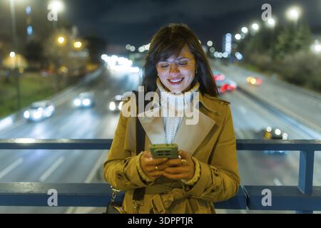 Young woman smiling and engaging with a mobile phone, standing on an urban bridge overlooking a busy highway with bokeh lights at night, embodying con Stock Photo