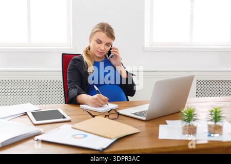Pregnant Woman Working at Desk While Talking on Phone and Taking Notes Stock Photo