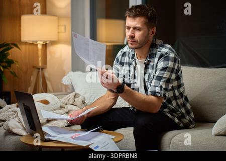 Man sitting on couch at home examining paper bills and financial documents with laptop on table. Concept of tracking personal expenses and managing mo Stock Photo