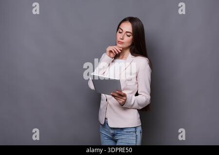 Young Woman in a Light Jacket Thinking While Using a Tablet Against a Gray Background Stock Photo