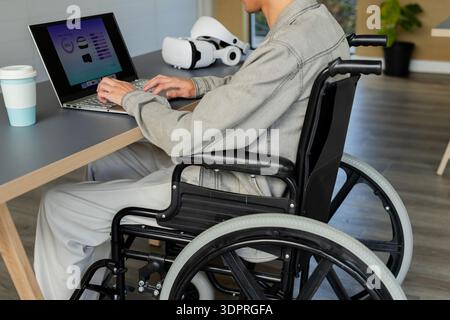 Laptop displaying data charts and VR headset sitting on wood office desk near manual wheelchair Stock Photo