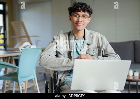 Man sitting in wheelchair using laptop in modern lounge wearing denim jacket and eyeglasses Stock Photo