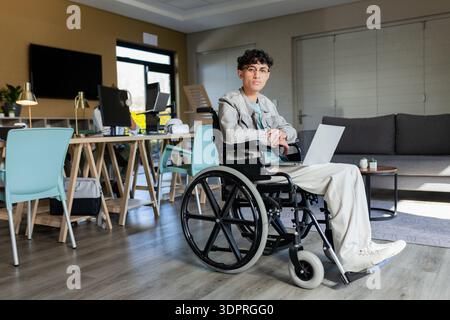 Asian man in wheelchair typing on laptop in modern office space with monitors and pastel chairs Stock Photo