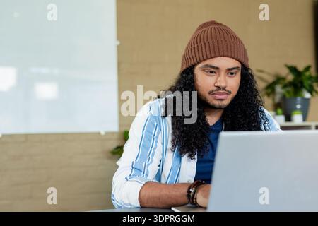 Indian man wearing brown beanie sitting at office desk working on laptop with whiteboard and plants Stock Photo