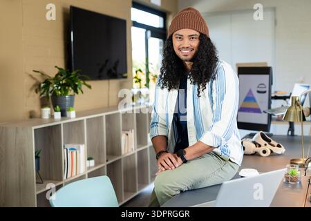 Asian man sitting on desk edge in open-plan office using laptop beside VR headset, copy space Stock Photo