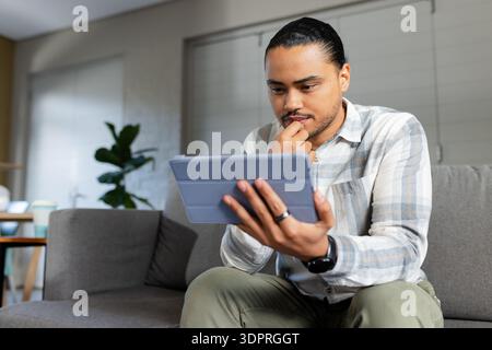 Casually dressed man leaning on gray couch in living room browsing tablet propping chin, copy space Stock Photo