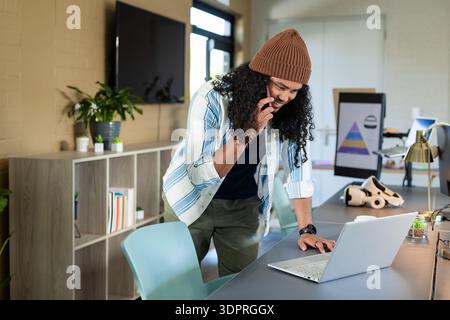 Hispanic mid adult man leaning on laptop and holding smartphone near VR headset at open-plan office Stock Photo