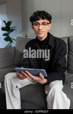 Asian man sitting on sofa holding tablet near potted plant wearing black jacket in living room Stock Photo