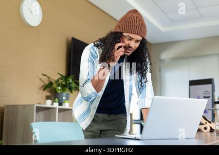 Mid adult man leaning forward at conference table, talking on smartphone and checking laptop screen Stock Photo