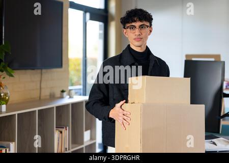 Male adult holding two stacked cardboard boxes in office with wall-mounted screen and desk monitor Stock Photo