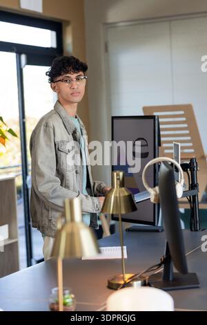 Male wearing denim jacket standing at office workspace typing on keyboard beside dual monitors Stock Photo