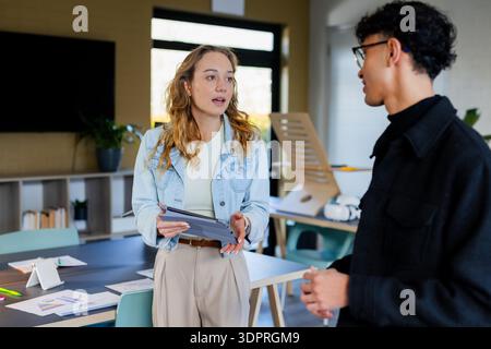 Diverse coworkers standing at table in office discussing project with tablet and charts Stock Photo