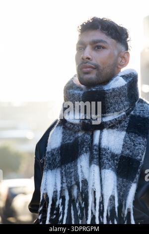 Man standing on backlit street wearing plaid scarf, dark jacket and shoulder strap, gazing right Stock Photo