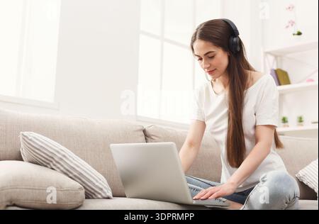 Young Woman Sits on Couch Using Laptop With Headphones in Bright Room During Day Stock Photo