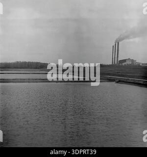 Donetsk region, Ukraine, USSR. 1960s-1970s. Panoramic industrial landscape of the Northern Donbas near the Donrybkombinat. In the foreground, the dark rippling water surface of large artificial fish farming ponds stretches out. In the background, a massive Soviet thermal power plant with three tall smoking chimneys dominates the cloudy horizon line. A dense forest belt runs along the left side. The symbiotic coexistence of heavy energy industry and aquaculture. Historic view of the peaceful Donbas before the war Stock Photo