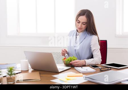 Office Worker Eating Salad While Using Laptop During Lunch Break in Bright Workspace Stock Photo