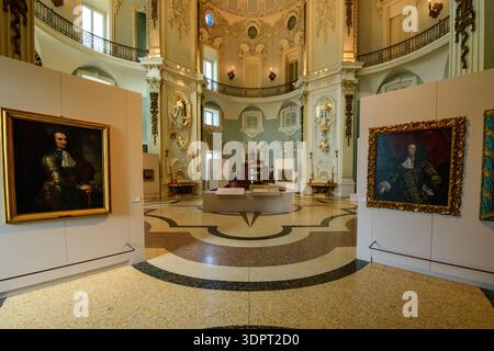 Isola Bella, Italy - September 30, 2020: The image shows a grand baroque hall inside Palais Borromee featuring high ceilings, ornate stucco decorations, balcony railings, large portrait paintings in gilded frames on display panels, and museum exhibits arranged in the center of the room. Stock Photo