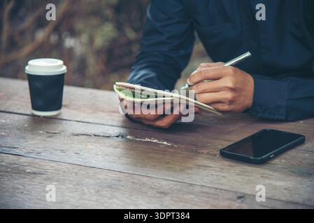 Man hands writing notebook diary with coffee cup and smartphone on wood desk. Close up man hands using pen sitting at wooden table outdoors lifestyle. Stock Photo