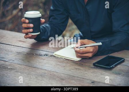 Man hands writing notebook diary with coffee cup and smartphone on wood desk. Close up man hands using pen sitting at wooden table outdoors lifestyle. Stock Photo