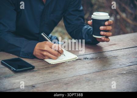 Man hands writing notebook diary with coffee cup and smartphone on wood desk. Close up man hands using pen sitting at wooden table outdoors lifestyle. Stock Photo