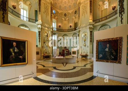Isola Bella, Italy - September 30, 2020: The image shows an ornate baroque hall inside Palais Borromee with high ceilings, decorative stucco, balcony railings, and framed portrait paintings on display panels, along with museum exhibits in the center of the room. Stock Photo