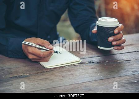 Man hands writing notebook diary with coffee cup and smartphone on wood desk. Close up man hands using pen sitting at wooden table outdoors lifestyle. Stock Photo