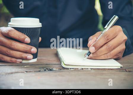 Man hands writing notebook diary with coffee cup and smartphone on wood desk. Close up man hands using pen sitting at wooden table outdoors lifestyle. Stock Photo