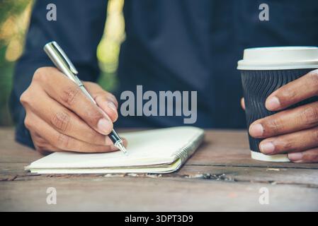 Man hands writing notebook diary with coffee cup and smartphone on wood desk. Close up man hands using pen sitting at wooden table outdoors lifestyle. Stock Photo