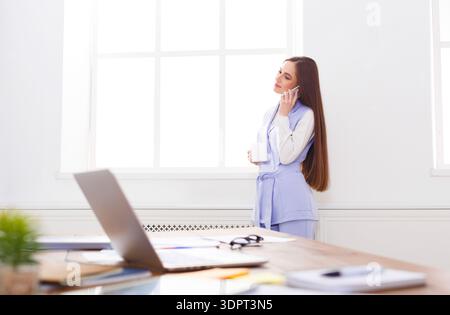 Woman Talks on the Phone While Holding a Cup in a Bright Office Space During the Day Stock Photo
