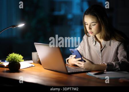 Serious businesswoman working in the night using phone and laptop at home Stock Photo