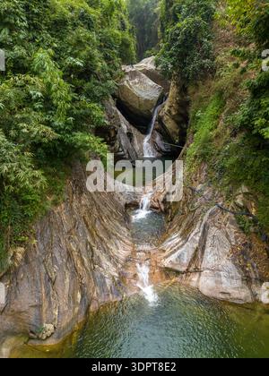 A small tiered waterfall flows through a narrow rock ravine into clear freshwater pools, surrounded by dense forest vegetation. Exposed rock surfaces Stock Photo
