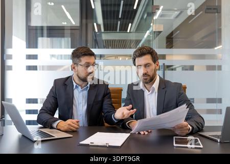 Two businessmen in suits collaborate over documents and laptops in a modern glass-walled meeting room, discussing strategy, analyzing data and planning next steps together Stock Photo