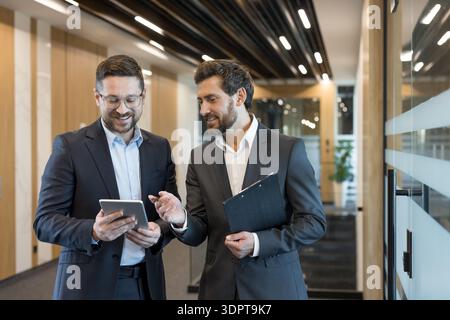 Two professional businessmen collaborate in a bright office corridor, reviewing a tablet and clipboard while smiling and discussing strategy, data and project plans Stock Photo