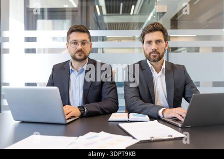 Two serious businessmen in suits sit at a desk with laptops and paperwork, facing the camera in a modern office, conveying corporate partnership, focused teamwork and communication Stock Photo