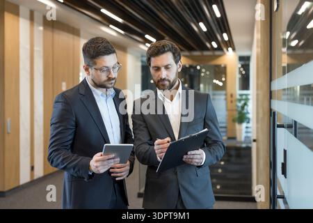 Two businessmen discussing business strategy and planning, collaborating and reviewing documents on a digital tablet and clipboard in a modern office hallway Stock Photo