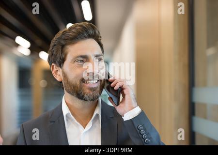 Smiling businessman wearing a suit communicating on a cell phone while walking in a bright office corridor, representing modern business communication, connectivity, and success Stock Photo