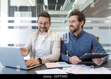 Two male business professionals are engaging in a collaborative meeting at a modern office, one pointing at a laptop screen while the other holds a clipboard, both smiling and discussing work Stock Photo