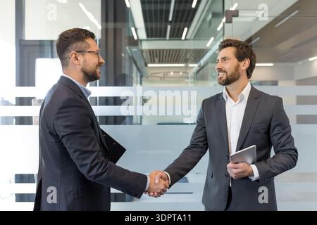 Two smiling businessmen in suits shaking hands in a modern office, symbolizing a successful partnership, agreement, collaboration, and a new professional deal Stock Photo